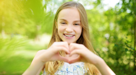 Cute preteen girl laughing and holding her hands in a heart shape on bright and sunny summer day. Cute child enjoying herself outdoors.