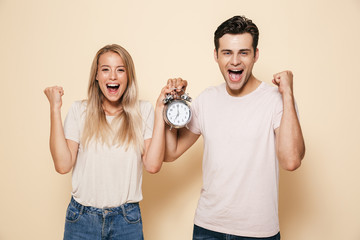Happy young loving couple holding alarm clock isolated make winner gesture.