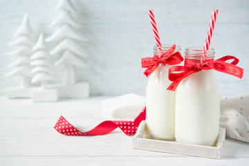 Festive bottles of milk with bows and straws on white wooden background with christmas trees...