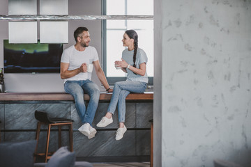 couple sitting on kitchen counter with cups of coffee in morning and looking at each other