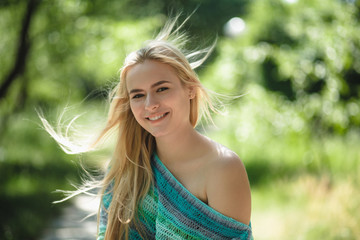 Portrait shot of good-looking beautiful woman outdoors. Smiling cheerful blonde lady wearing light blue sweater with one of her shoulders showing outside in park.