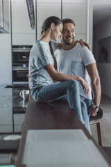 side view of attractive woman sitting on kitchen counter and embracing boyfriend at home