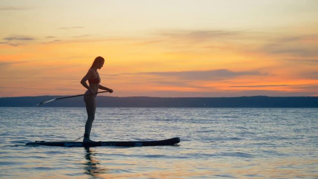 Slim attractive woman is moving on her paddleboard across the sunset waters