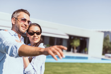 selective focus of adult man in sunglasses pointing by finger to girlfriend standing near on green lawn at country house