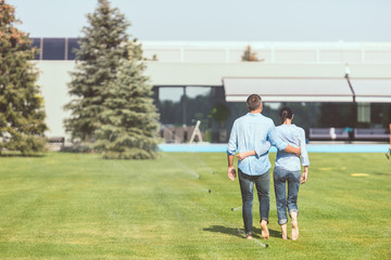 rear view of couple embracing each other and walking on lawn near country house