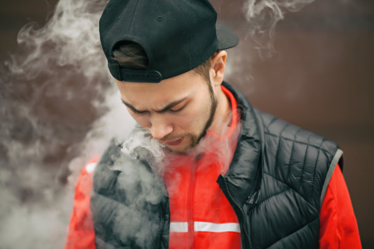 Vape teenager. Portrait of a handsome young white man in black cap vaping an electronic cigarette opposite the modern background in autumn.
