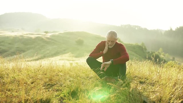Senior man runner sitting on meadow outdoors at sunrise in nature, using smartwatch.