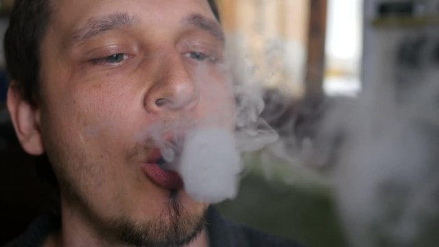 Man smokes electronic cigarette at home exhaling smoke rings closeup