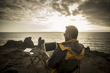 man traveler working during the sunset on a cliff with the ocean and the sun in front of him, freedom from office doing business everywhere like a digital nomad enjoying freedom 