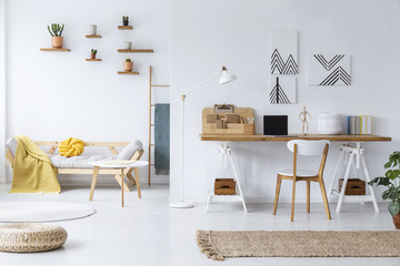 Chair at wooden desk and pouf in white open space interior with posters and table near sofa. Real...