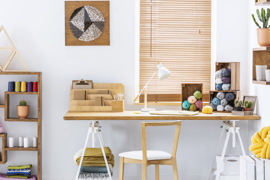 Wooden Chair At Desk With Lamp And Colorful Yarns In Home Office Interior With Poster. Real Photo