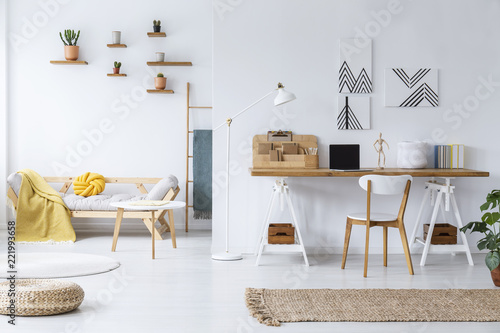 Chair at wooden desk and pouf in white open space interior with posters and table near sofa. Real photo