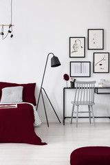 Real photo of an elegant bedroom interior with burgundy accents, lamp, chair and graphics collection