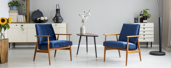 Fototapeta premium Real photo of bright living room interior with two royal blue armchairs, end table with flowers and cupboard with books