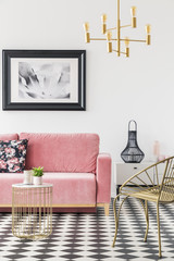 Gold armchair near table and pink couch in living room interior with lamp and poster. Real photo