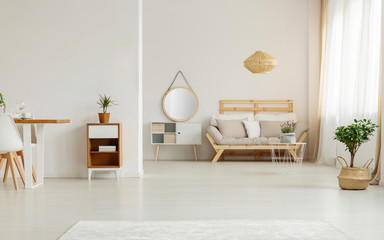 Mirror on white cabinet next to beige wooden sofa in living room interior with plants. Real photo