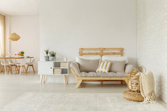 Blanket in basket in front of beige wooden sofa in white flat interior with plants on cabinet. Real photo