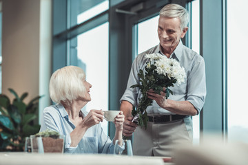 Here I am. Cheerful mature man keeping smile on his face while presenting flowers to his wife