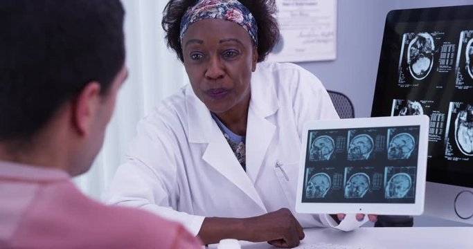 Portrait of mid aged African doctor using electronic tablet to show patient ct-scans of his brain. Close up of female doctor explaining to young latino patient results from his mri