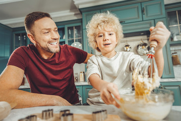 Baking mix. Blonde-haired curly cute son holding bowl with baking mix for cookies making them with...