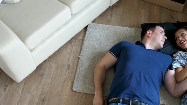 Husband and wife lying down on the floor of their new house. They are surrounded by cardboard boxes. The man is showing the keys to their new appartment to the woman