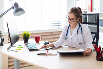 A young girl in a white robe is sitting at a computer desk, holding a pencil in her hand and...