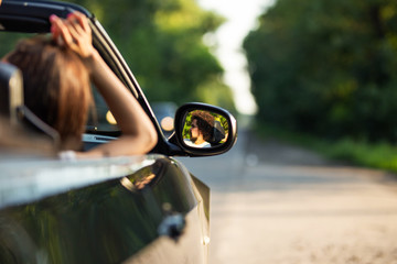 Black cabriolet on the road on a sunny day. In the side mirror is reflection of the dark-haired young man with beard.