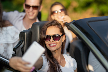 Attractive young brunette woman in sunglasses dressed in a white t-shirt sits with friends in a black cabriolet make a selfie on a sunny day.