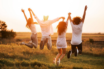 Fototapeta premium Dwóch młodych chłopaków i dwie dziewczyny trzymają rękę i skaczą w polu w letni dzień. Widok z tyłu.