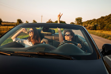 Two charming dark-haired young women in sunglasses are sitting in a black cabriolet on a sunny day.