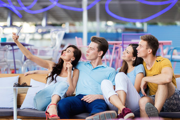 A company of good-looking friends is laughing and sitting on the bench with cushions and making selfie in the nice summer cafe. Entertainment, having good time. Friendship.