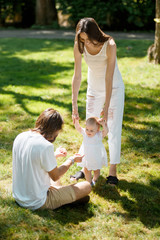 Fototapeta premium Exciting mother and father are teaching their little daughter wearing white dress how to make her first steps.