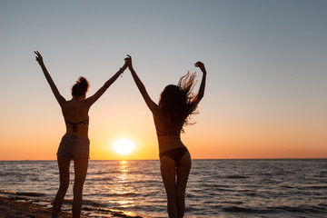 Two slim dark-haired girls in swimsuits hold hands up near the sea on the sunset