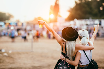 Two female friends drinking beer and having fun at music festival. Back view - obrazy, fototapety, plakaty