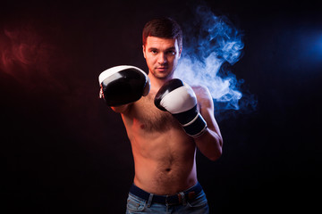 Fototapeta na wymiar Studio portrait of a muscular boxer in professional gloves of European appearance with light bristles and hair on his chest. Smoke in the background is illuminated in blue and red