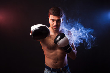 Studio portrait of a muscular boxer in professional gloves of European appearance with light bristles and hair on his chest. Smoke in the background is illuminated in blue and red