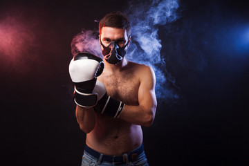 Studio portrait of a muscular boxer in professional gloves of European appearance with light bristles and hair on his chest. Smoke in the background is illuminated in blue and red