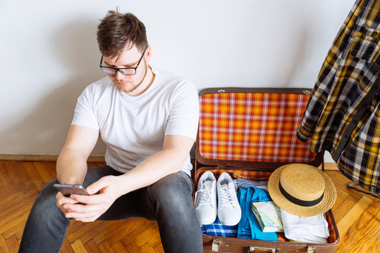 Young Man Sit On Floor Checking List On Mobile. Valise Packing For Trip.travel Concept. Copy Space.