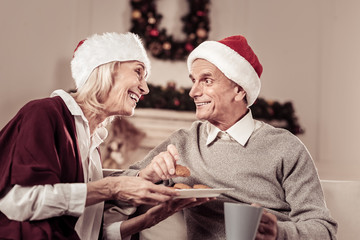 Holyday cakes. Joyful senior pleasant couple in red hats spending time together at home smiling and eating cakes.