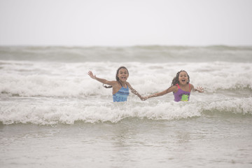 Two young pre-teen mixed race female sisters wearing swimwear screaming and having fun and playing in the crashing waves 