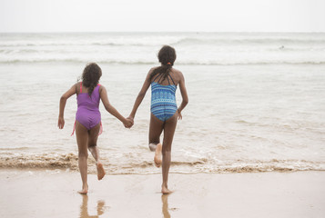 Fototapeta na wymiar Rear view of two mixed-race sisters running into the water playing and laughing having fun on the beach on a bright tropical summer holiday