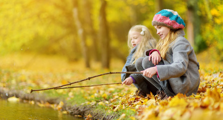 Cute little girls playing by the water on beautiful autumn day. Happy children having fun in autumn park. - obrazy, fototapety, plakaty