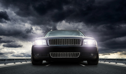 Plakat A powerful car with lights on stands on the highway at sunset against a dramatic sky.