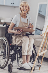 Artist in wheelchair. Blonde-haired smiling artist feeling inspired while sitting in wheelchair holding color palette for drawing