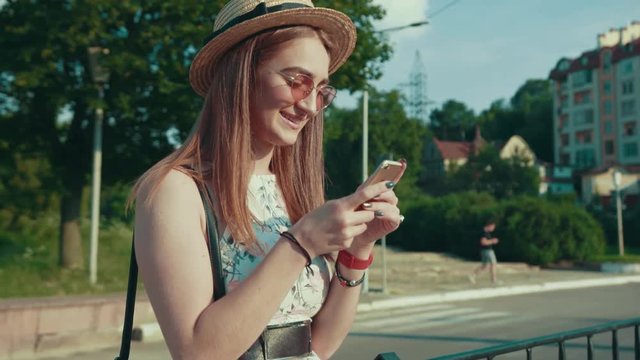 Cute smiling young woman in trendy summer straw hat stands outdoors on a bus stop actively using her smartphone. Being online, social networks, technologies concept. Female portrait
