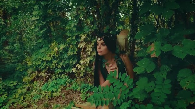 A forest nymph with long dark hair walks with a white owl. A girl in an unusual green dress with a deep neckline and bare legs. Background wild nature