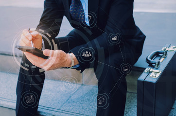 hand of business man wearing modern black suit using mobile smart phone and briefcase sitting on walkway in city with graphic network diagram, technology communication, internet and financial concept