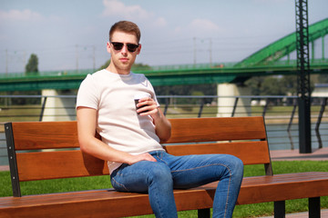 Handsome boy wearing sunglasses, sitting on a bench on a pier and drinking coffee to go. Taking a break from city rush. Background scenery bridge, river, pier, path along the river and green grass.