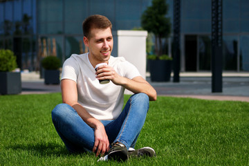 Young handsome guy drinking coffee to go, man sitting on grass and enjoying a nice sunny day by modern building side. Male model photo-shoot, urban fashion, hipster lifestyle. White T-shirt and jeans.