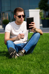 Young handsome guy with sunglasses using tablet and headphones, drinking coffee to go, sitting on grass and enjoying nice sunny day by modern building. Male model photo-shoot, urban fashion lifestyle.